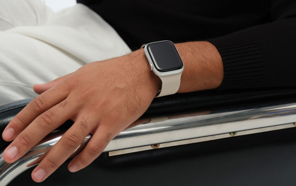 Osoba siedzi na krześle, nosząc biało-czarny pasek do zegarka Apple Watch na nadgarstku. Zegarek ma prostokątną tarczę z czarnym ekranem.