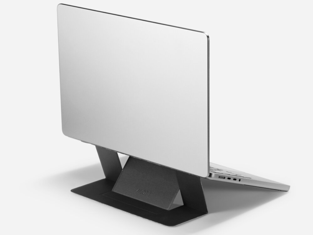 Na obrazie widoczny jest laptop umieszczony na czarnej podstawce, która unosi go pod kątem. Na podstawce znajduje się napis \'MOFT\'.