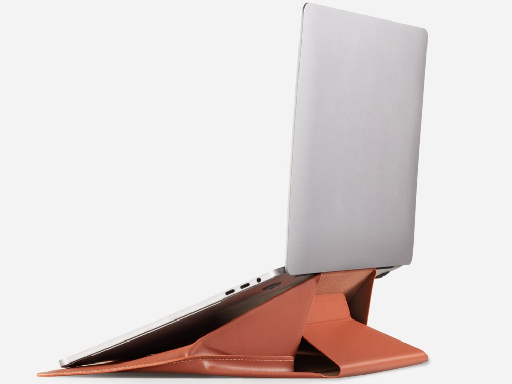 Na obrazie widoczna jest brązowa torba na laptopa, która pełni funkcję podstawki, z umieszczonym na niej otwartym laptopem. Torba ma nowoczesny design i jest ustawiona w sposób umożliwiający podparcie urządzenia.