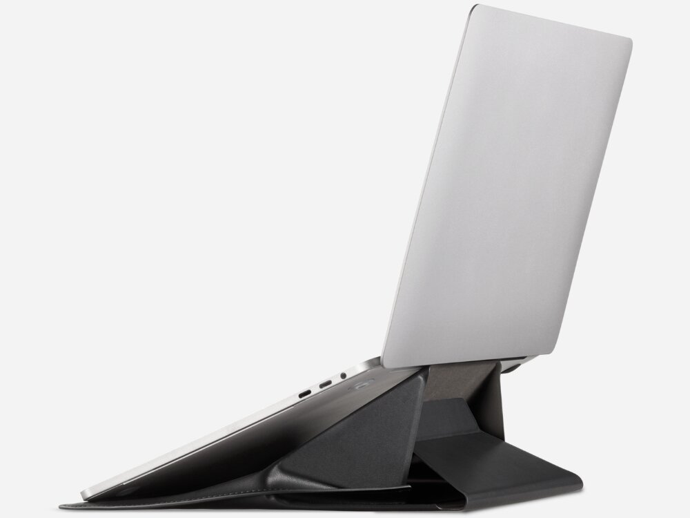 Na obrazie widoczny jest laptop umieszczony na czarnej podstawce, która unosi go pod kątem. Podstawka ma geometryczny kształt i jest wykonana z materiału przypominającego skórę.