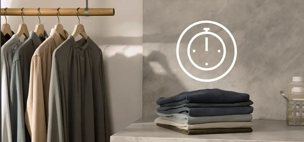 Na obrazie widać kilka ubrań wiszących na drewnianym wieszaku oraz stos złożonych ubrań na blacie. Obok znajduje się symbol zegara na ścianie.