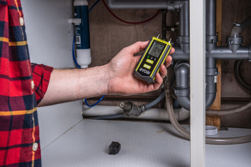 Osoba trzyma miernik wilgotności z wyświetlaczem pokazującym \'0.4%\', na którym widoczne jest logo \'RYOBI\'. W tle znajdują się rury i elementy instalacji wodnej pod zlewem.