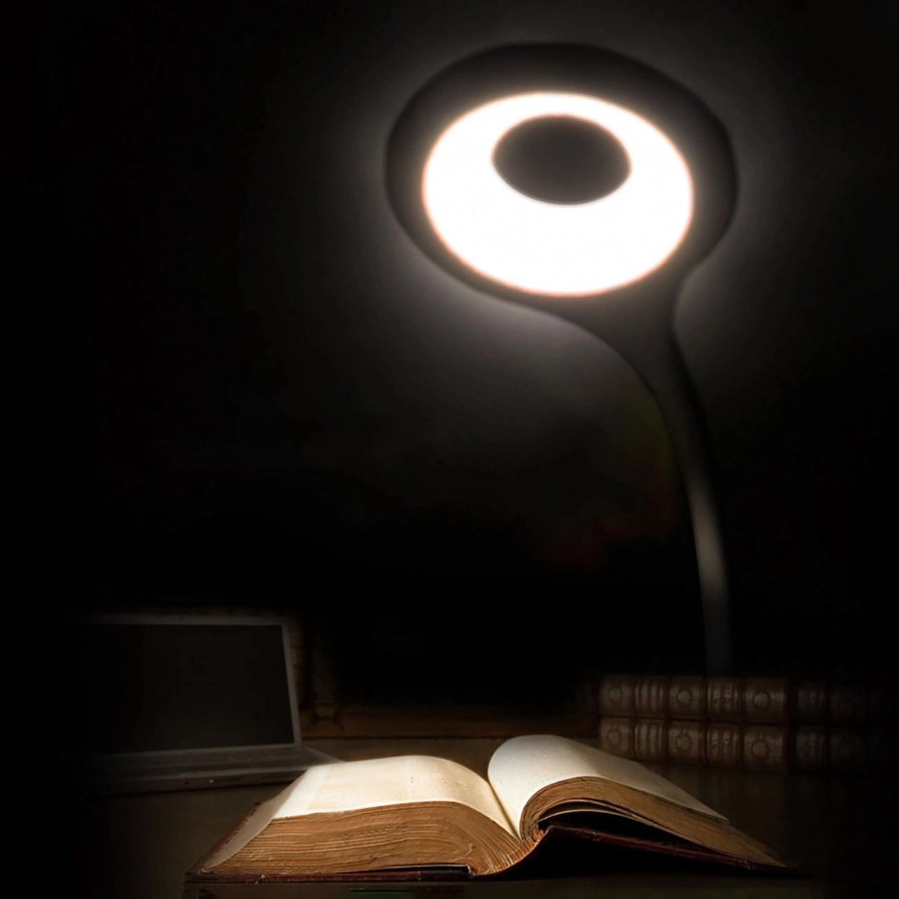 Na obrazie widoczna jest okrągła lampka LED emitująca jasne światło, oświetlająca otwartą książkę na ciemnym tle. W tle znajduje się zamknięty laptop i stos książek.