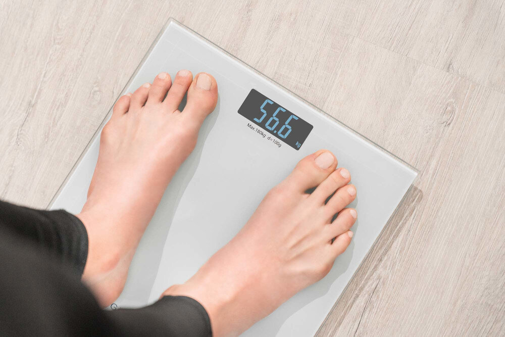 Osoba stoi na szklanej wadze łazienkowej, która pokazuje wynik 56.6 kg. Na wyświetlaczu widoczny jest również napis \'Max 180kg d=100g\'.
