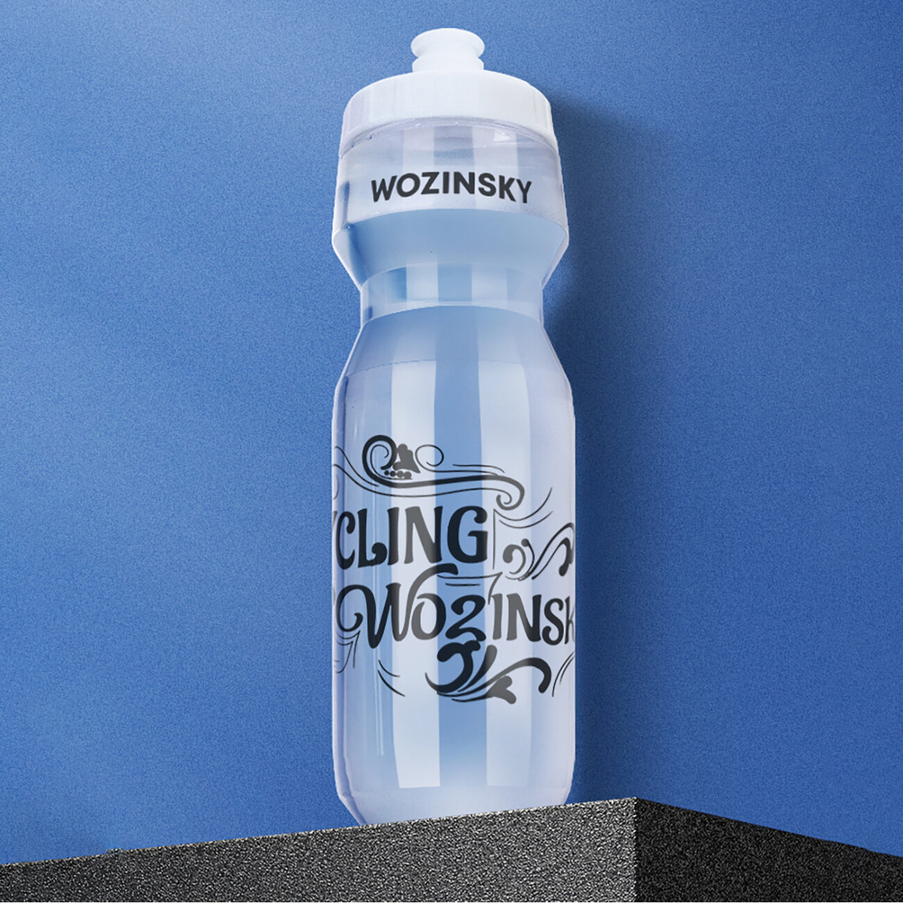 Przezroczysty bidon rowerowy z białą zakrętką, na którym widnieje napis \'WOZINSKY\' oraz ozdobny napis \'CLING WOZINSKY\'. Bidon stoi na ciemnej powierzchni, a tło jest niebieskie.