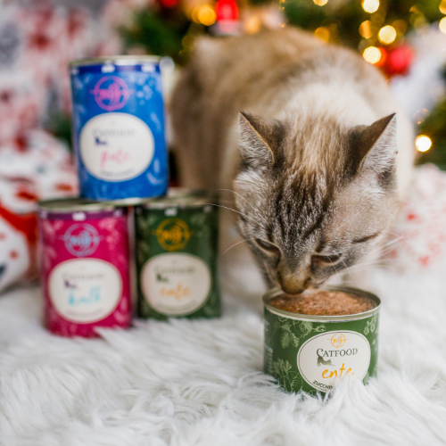 Kot wącha otwartą puszkę karmy o nazwie \'Catfood entree\'. W tle widoczne są cztery zamknięte puszki karmy w różnych kolorach.