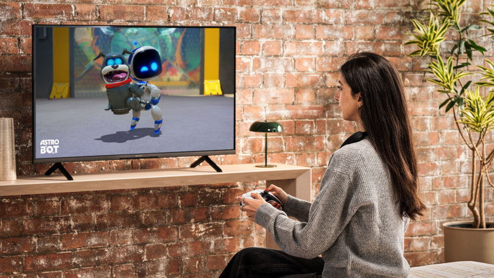Osoba siedzi przed telewizorem, na którym wyświetlana jest gra z postacią robota i napisem \'ASTRO BOT\'. W tle widoczna jest ceglana ściana i roślina w doniczce.