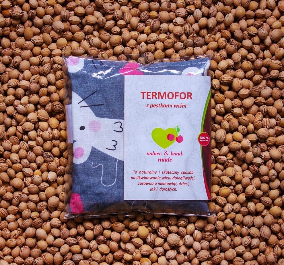 Na obrazie widoczny jest termofor z pestkami wiśni w opakowaniu, leżący na tle pestek. Na opakowaniu znajduje się tekst: \'TERMOFOR z pestkami wiśni nature & hand made To naturalny i skuteczny sposób na likwidowanie wielu dolegliwości, zarówno u niemowląt, dzieci, jak i dorosłych.\'