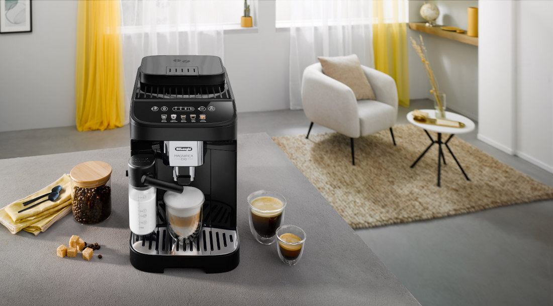 Ekspres do kawy z logo \'DELONGHI Magnifica\'. Obok znajdują się filiżanki z kawą i słoik z ziarnami kawy.