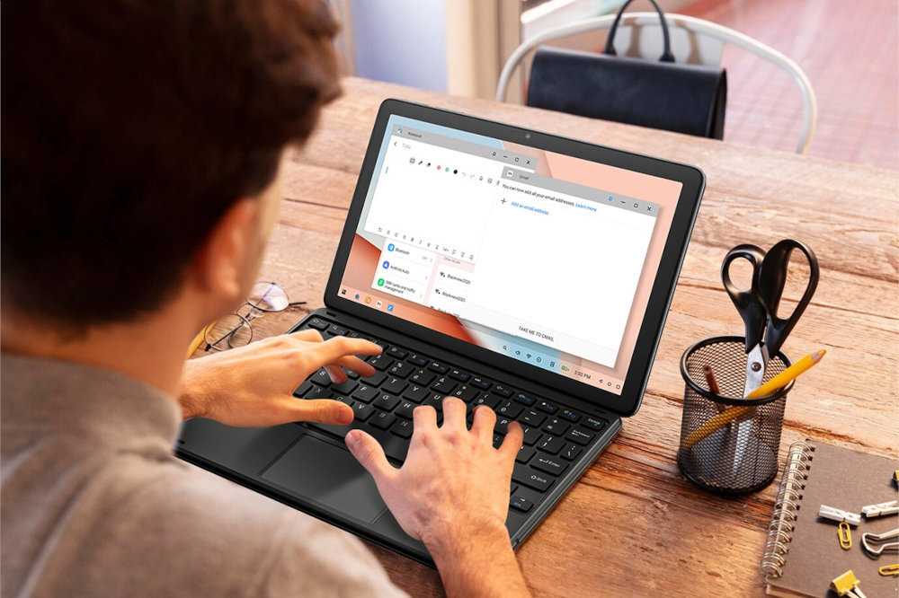 Osoba używa tabletu z klawiaturą na drewnianym stole. Obok tabletu znajduje się pojemnik z przyborami do pisania i notes.