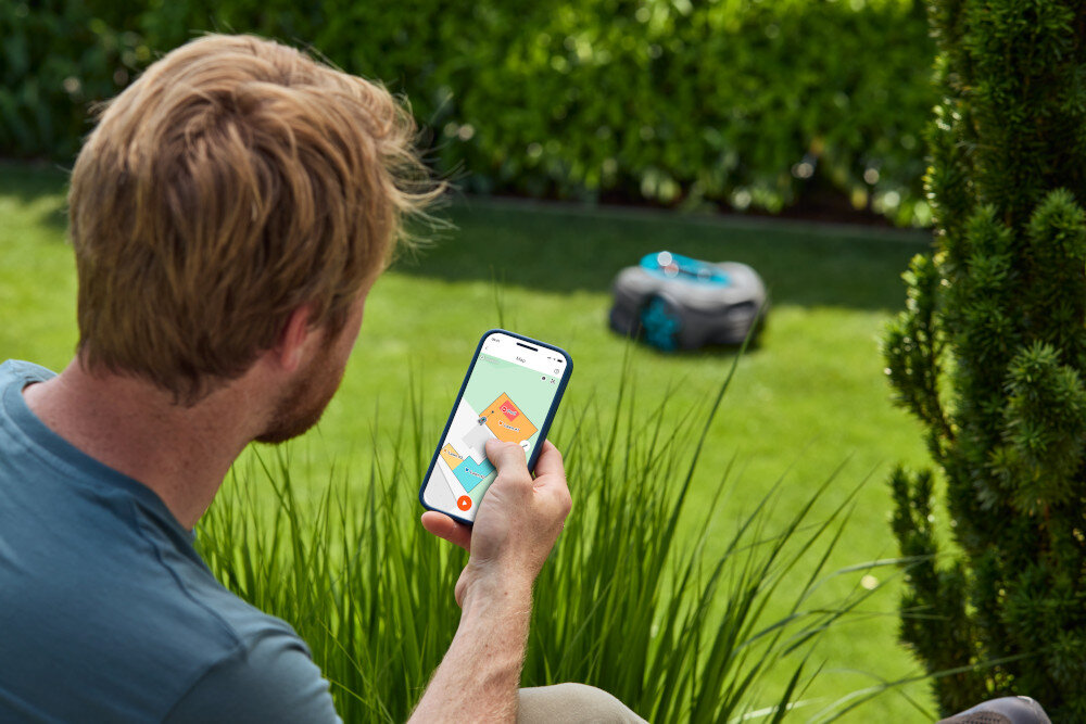 Osoba trzyma smartfon z aplikacją pokazującą mapę trawnika. W tle na trawie znajduje się robot koszący.