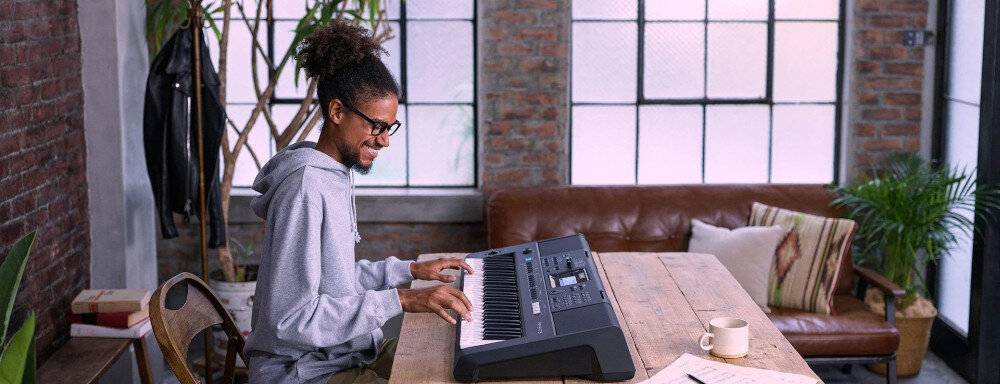 Osoba gra na czarnym keyboardzie YAMAHA PSR-E473, siedząc przy drewnianym stole w jasnym pomieszczeniu z dużymi oknami. W tle znajduje się brązowa sofa i rośliny doniczkowe.