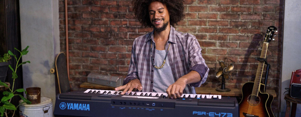 Osoba gra na keyboardzie YAMAHA PSR-E473 Czarny, stojącym na tle ceglanej ściany. Obok znajduje się gitara akustyczna i wentylator.