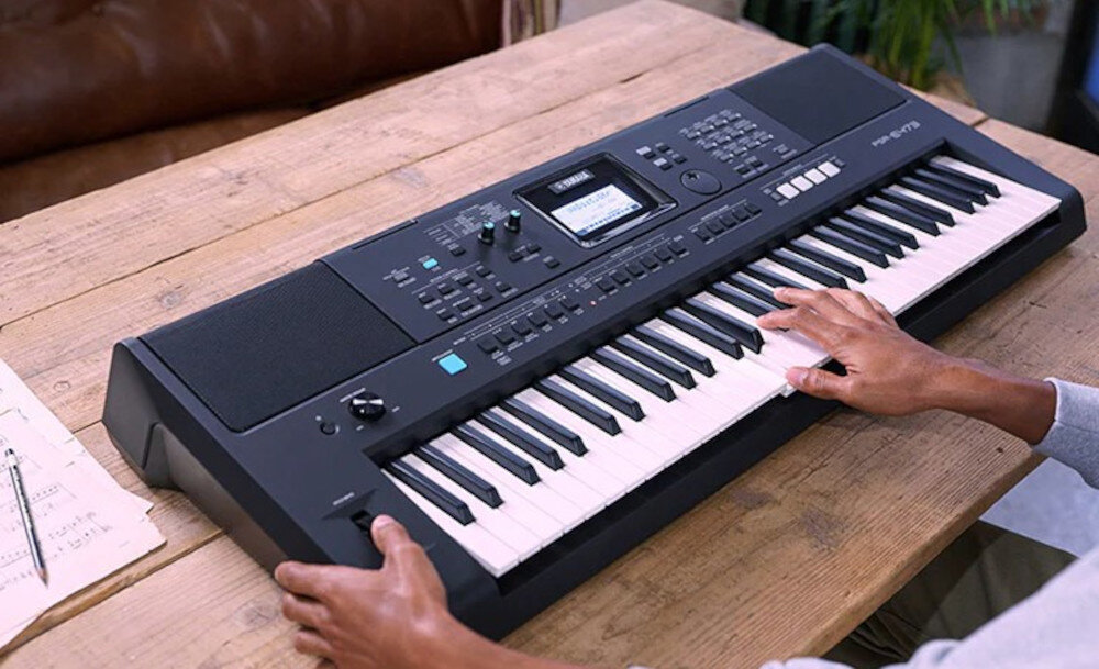 Osoba gra na czarnym keyboardzie z wyświetlaczem i wieloma przyciskami na drewnianym stole. Obok leży kartka z nutami.