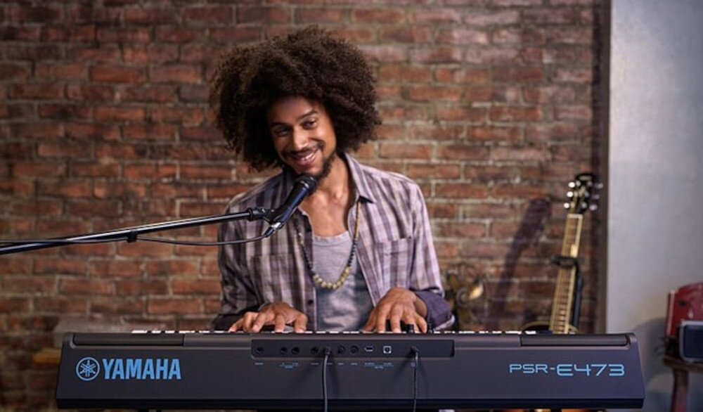 Osoba gra na keyboardzie YAMAHA PSR-E473 Czarny, na tle ceglanej ściany. Obok keyboardu widoczne są gitary.