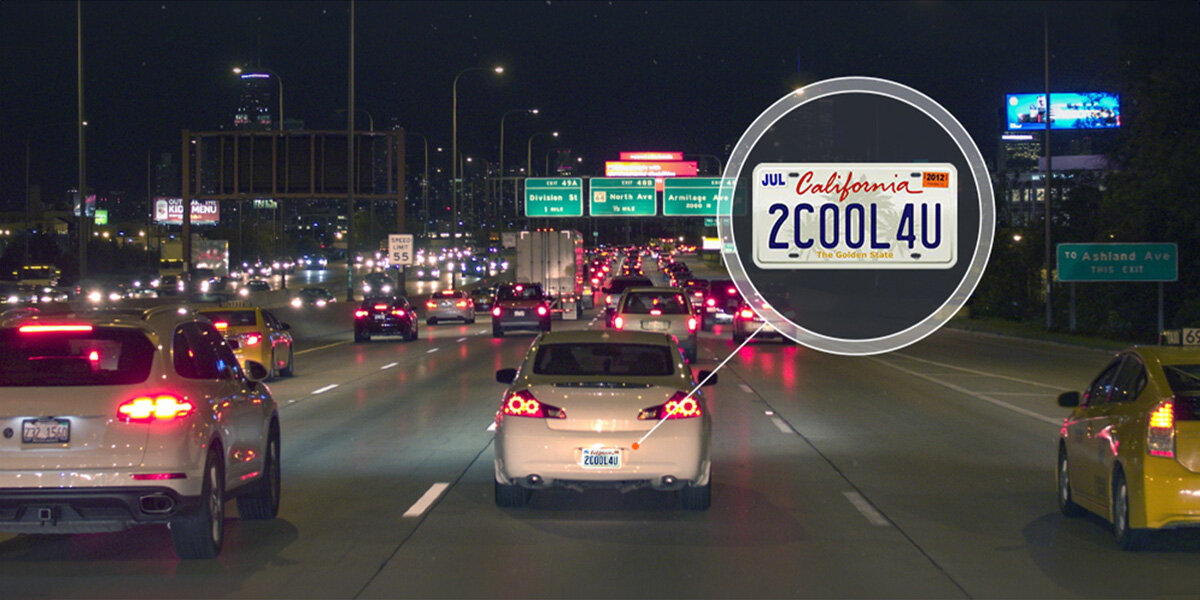 Nocna scena na autostradzie z dużym ruchem samochodowym. Powiększenie tablicy rejestracyjnej jednego z pojazdów pokazuje tekst \'California 2COOL4U\'.