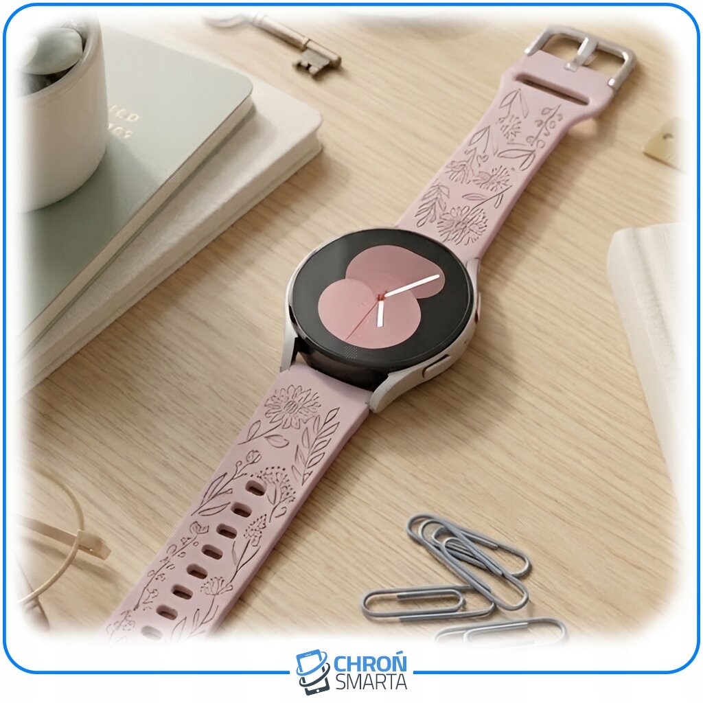 Smartwatch z różowym paskiem ozdobionym wzorami kwiatowymi leży na drewnianym blacie. W tle widoczne są książki, spinacze i klucz, a na dole logo \'CHROŃ SMARTA\'.