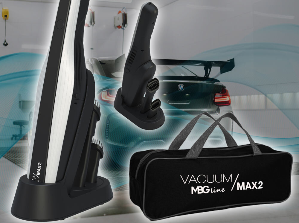Na obrazie widoczny jest odkurzacz ręczny w kolorze czarnym z białymi elementami, stojący na podstawce. Obok znajduje się czarna torba z napisem \'VACUUM MBG line MAX2\'.