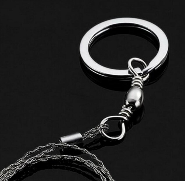 Na czarnym tle widoczna jest metalowa piła ręczna strunowa z okrągłym uchwytem i złączem. Struna jest spleciona z cienkich metalowych drutów.