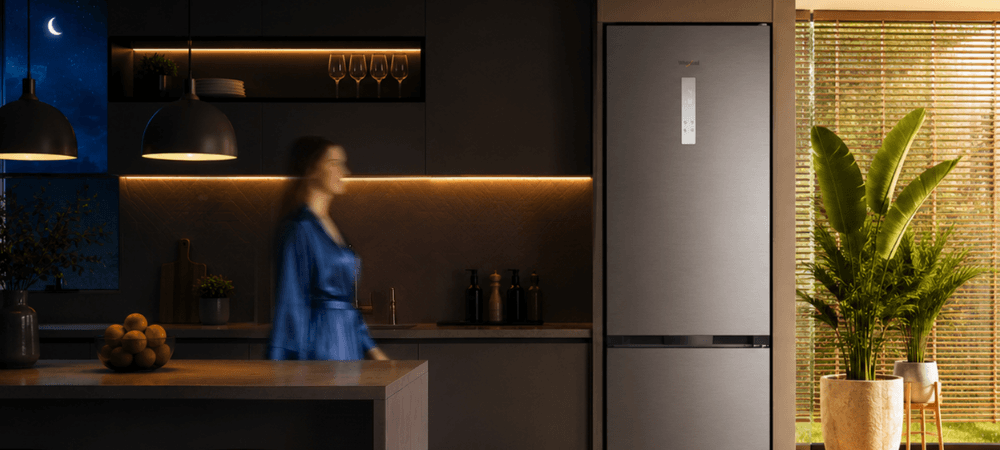 LODÓWKA WHIRLPOOL WHK 26412 X5EA2 Lodówka Whirlpool stoi w kuchennej zabudowie obok dużej rośliny doniczkowej. Po lewej stronie widoczna jest rozmyta sylwetka osoby, a w lewym górnym rogu znajduje się ikona księżyca wykończenie inox elegancki wygląd ponadczasowy design