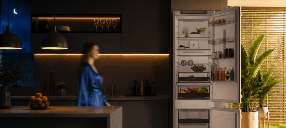 LODÓWKA WHIRLPOOL WHK2 5293 B5E Lodówka Whirlpool stoi w kuchennej zabudowie obok dużej rośliny doniczkowej. Po lewej stronie widoczna jest rozmyta sylwetka osoby, a w lewym górnym rogu znajduje się ikona księżyca wykończenie inox elegancki wygląd ponadczasowy design