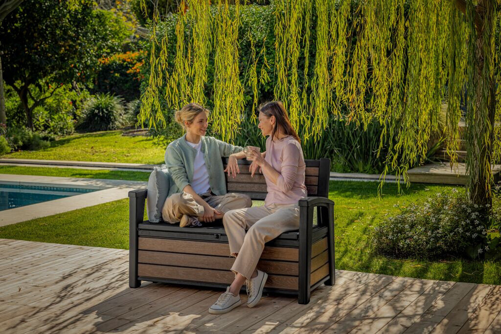 Dwie osoby siedzą na brązowej ławce ogrodowej KETER Deco Garden Bench, otoczonej zielenią i drzewami. W tle widoczny jest basen i zadbany trawnik.