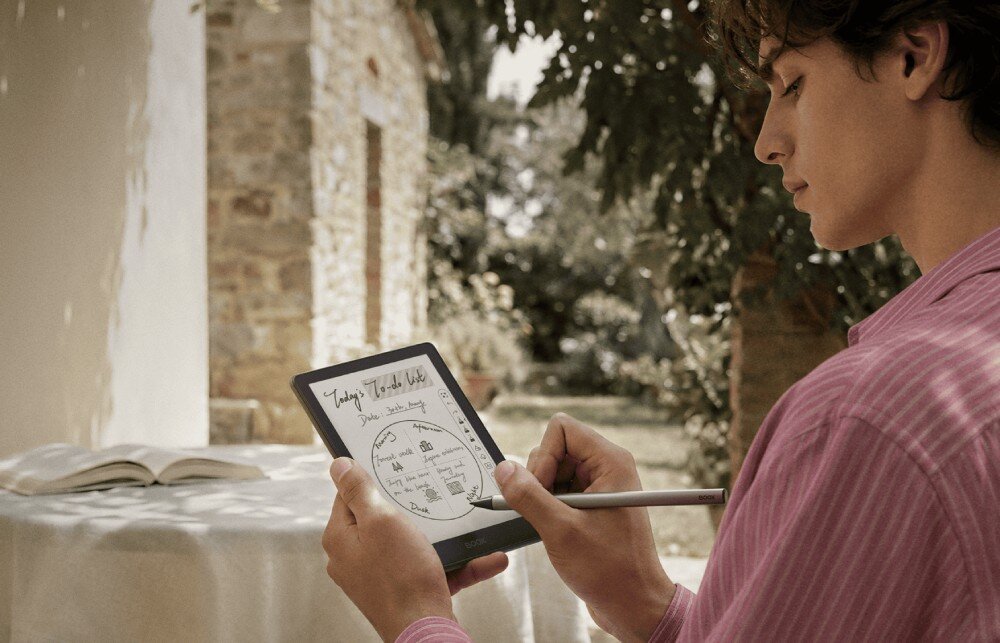 Czytnik e-booków ONYX Boox Go Mężczyzna w różowej koszuli rysuje okrągły diagram rysikiem na ekranie czytnika na zewnątrz na tle kamiennego domu i zieleni, rysik, obsługa