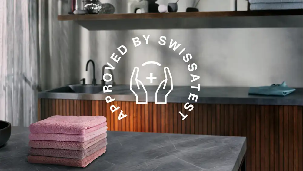 Na blacie kuchennym leży stos różowych ręczników, a w tle znajduje się zlew. Na obrazie widnieje napis \'APPROVED BY SWISSTEST\' z symbolem dłoni i plusem.