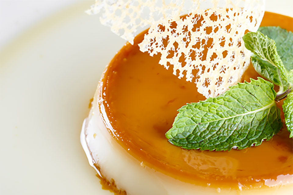 Dzięki funkcji Home Dessert każdy przepis wyjdzie smacznie Na obrazie widoczny jest deser z karmelowym wierzchem, ozdobiony listkami mięty i delikatną, ażurową dekoracją. Deser znajduje się na jasnym talerzu.