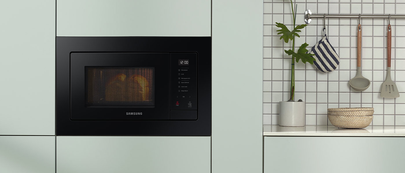 Kuchenka mikrofalowa do zabudowy Samsung zapewnia funkcjonalność i bezpieczeństwo w trakcie działania Czarna kuchenka mikrofalowa wbudowana w jasną szafkę kuchenną, z wyświetlaczem pokazującym godzinę 12:01. Obok znajduje się ściana z białych płytek, na której wiszą przybory kuchenne i roślina w doniczce.