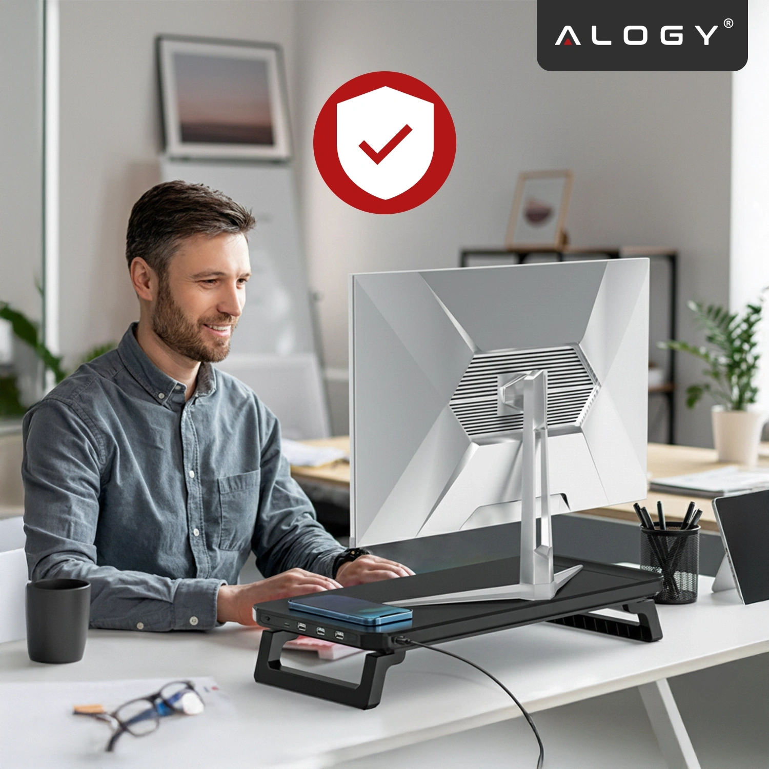 Na biurku znajduje się czarna podstawka pod monitor z oświetleniem RGB i portami USB, na której stoi monitor. W tle widoczny jest czerwony symbol tarczy z białym haczykiem oraz logo \'ALOGY\'.