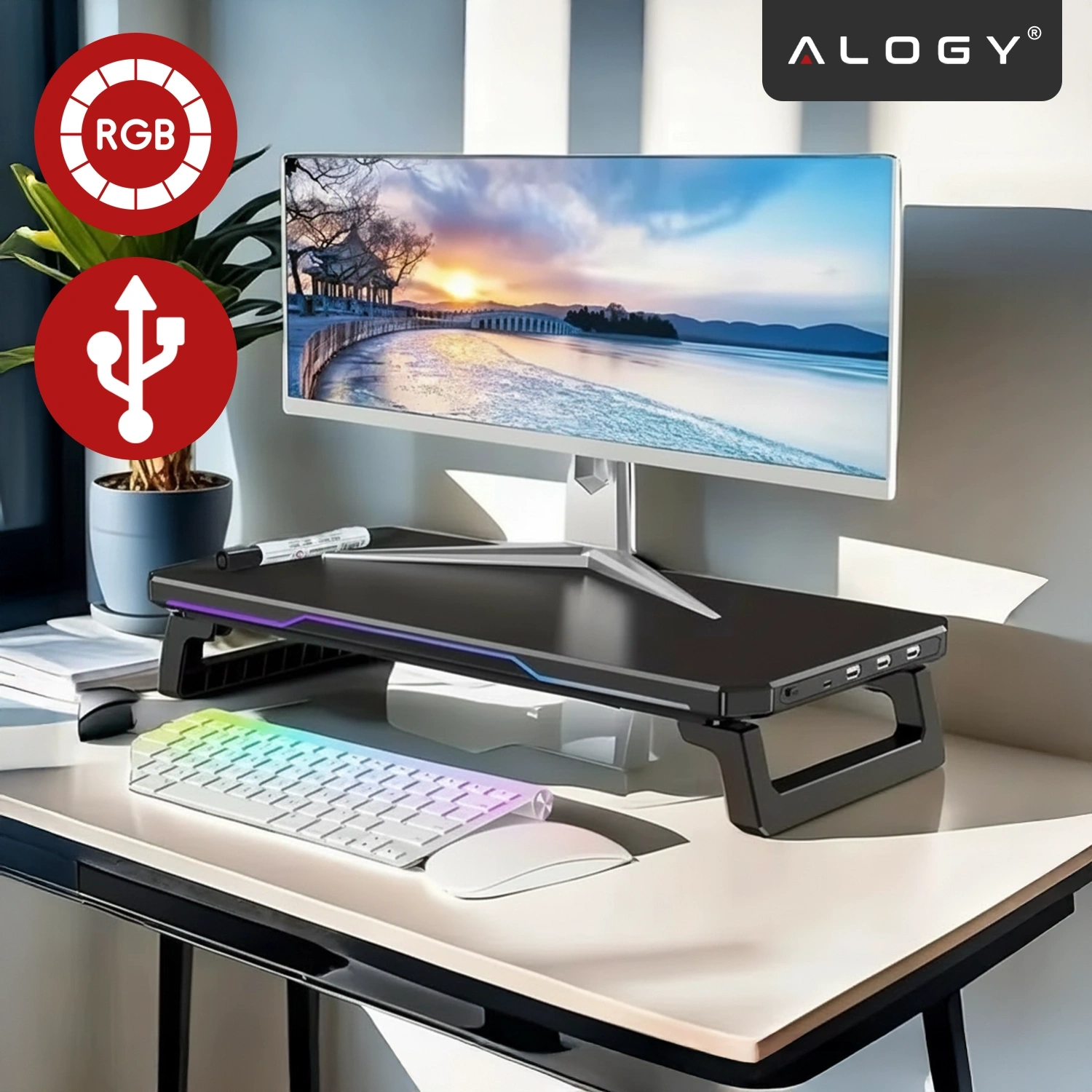 Na biurku znajduje się czarna podstawka pod monitor z oświetleniem RGB i portami USB, na której stoi monitor z krajobrazem. Obok widoczna jest klawiatura z podświetleniem, myszka oraz logo \'ALOGY\'.