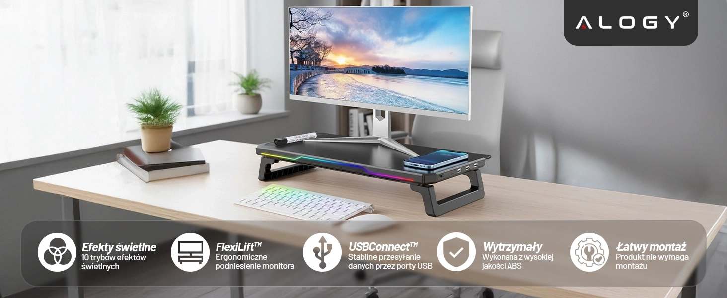 Na biurku znajduje się podstawka pod monitor z oświetleniem RGB, na której stoi monitor. W prawym górnym rogu widoczne jest logo \'ALOGY\'.