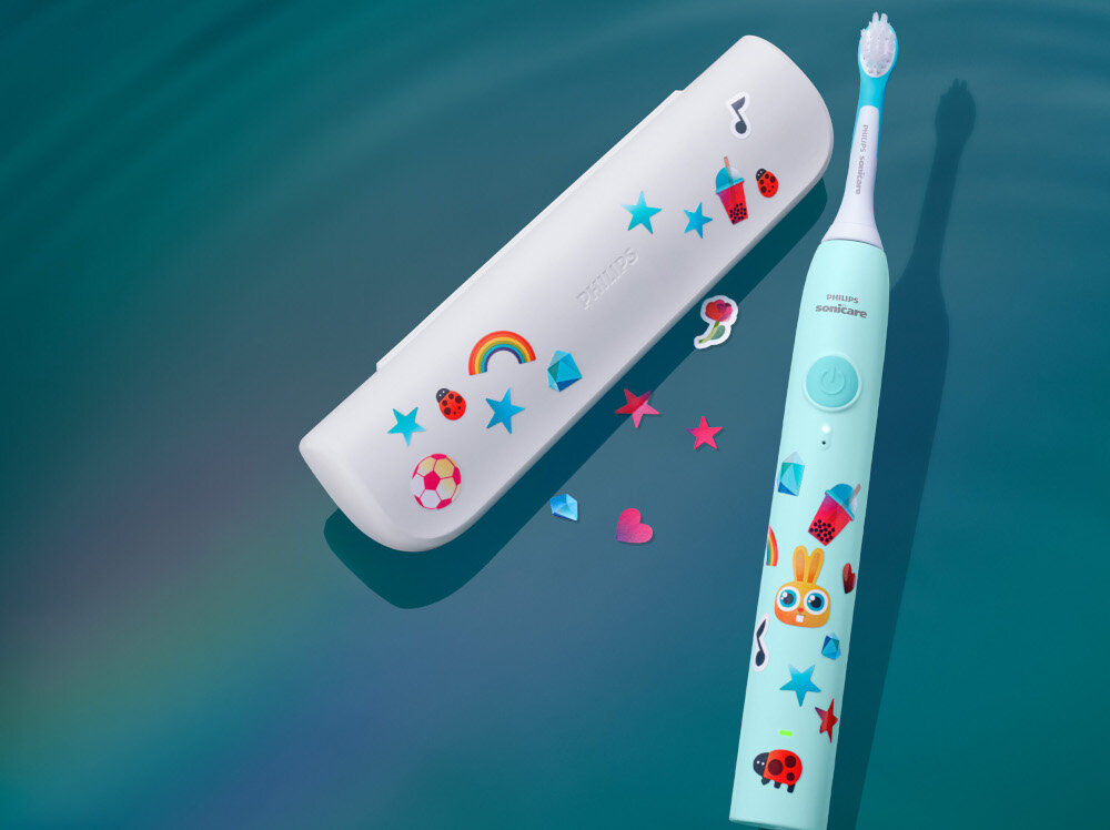 Szczoteczka soniczna w turkusowym kolorze z kolorowymi naklejkami, obok białe etui również ozdobione naklejkami. Na szczoteczce widoczny napis \'Philips Sonicare\'.