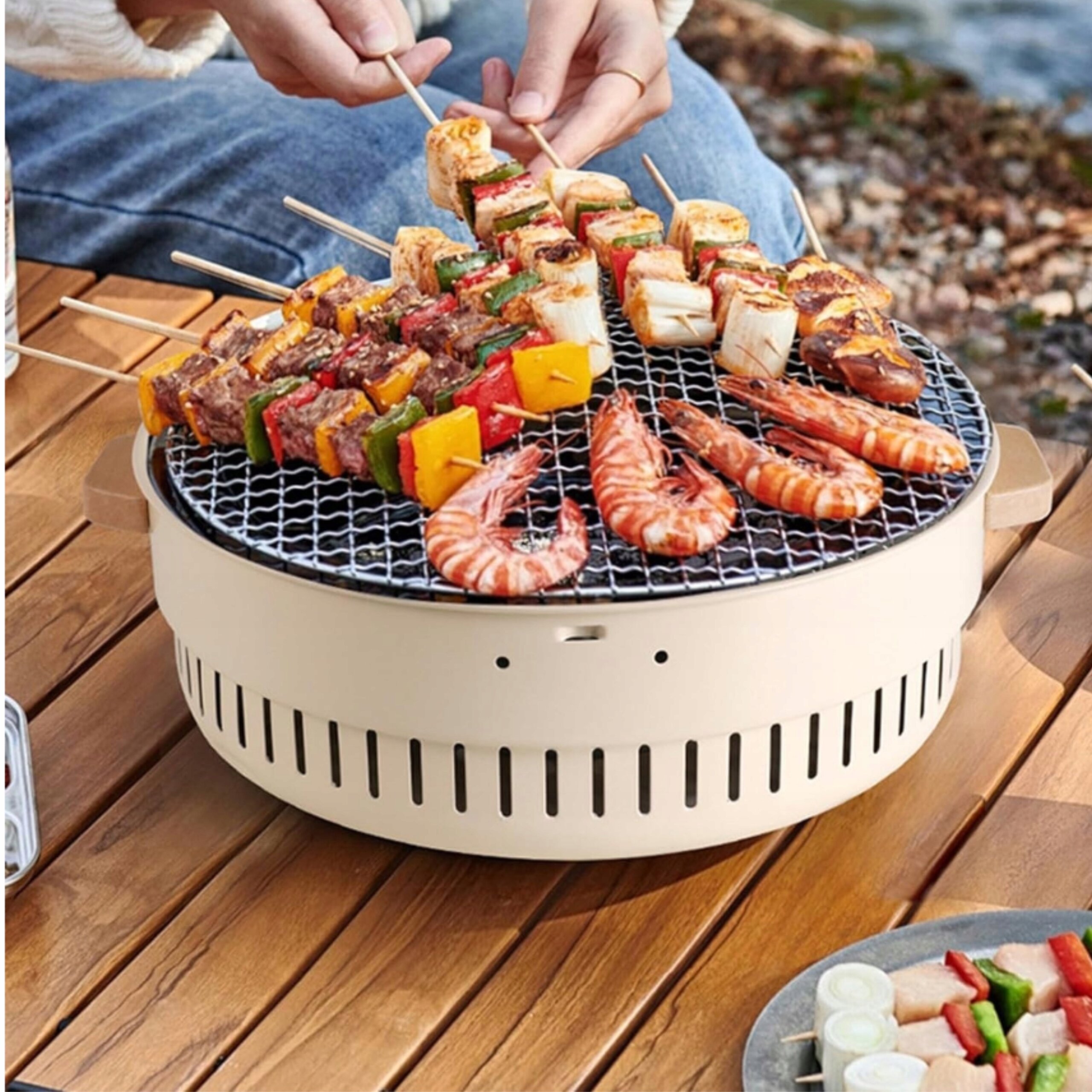 Na drewnianym stole znajduje się przenośny grill z rusztem, na którym grillowane są szaszłyki i krewetki. W tle widać osobę przygotowującą jedzenie.