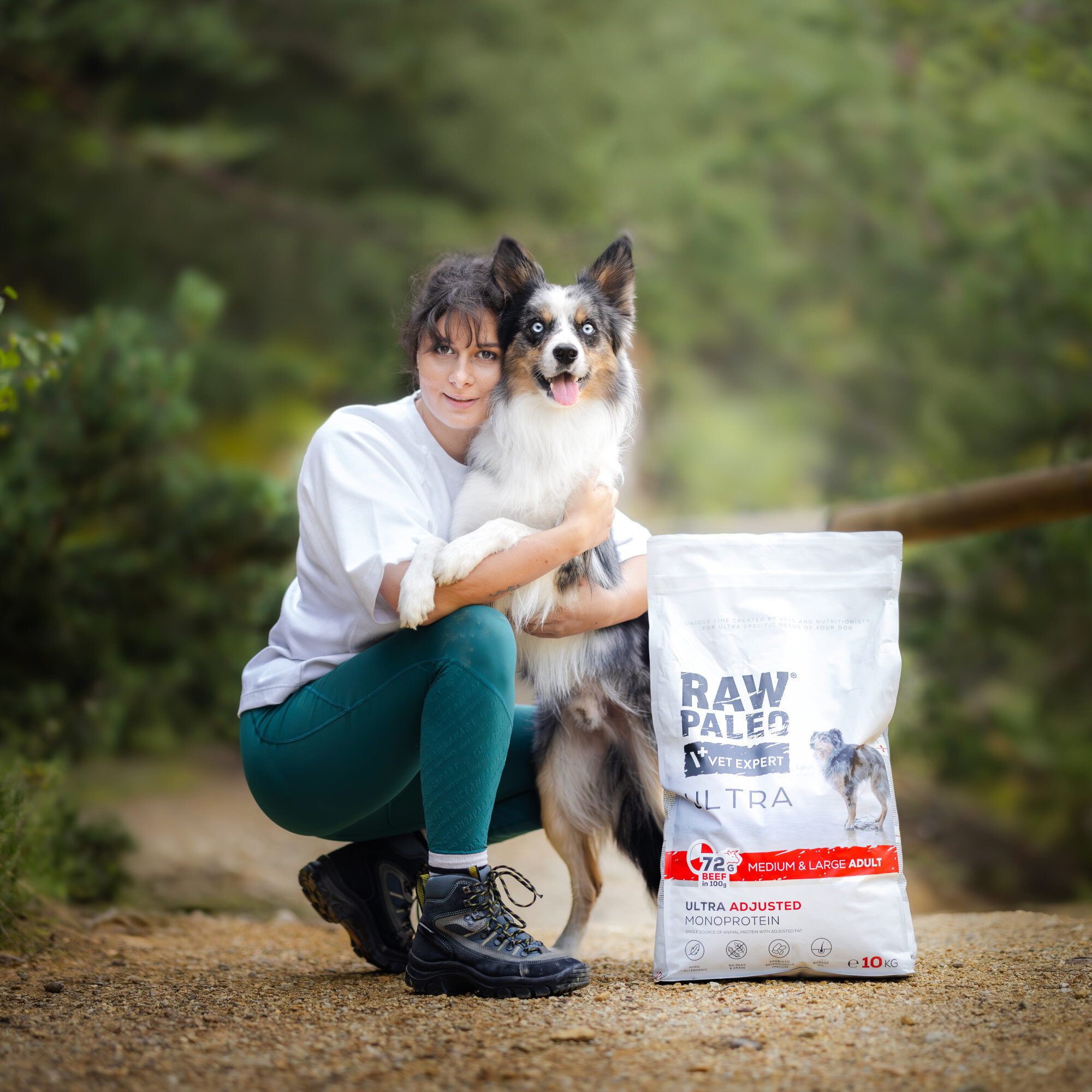 Osoba klęczy obok psa, trzymając go za łapy, na tle zielonego krajobrazu. Obok znajduje się worek z karmą dla psów z napisem \'RAW PALEO VET EXPERT ULTRA MEDIUM & LARGE ADULT\'.
