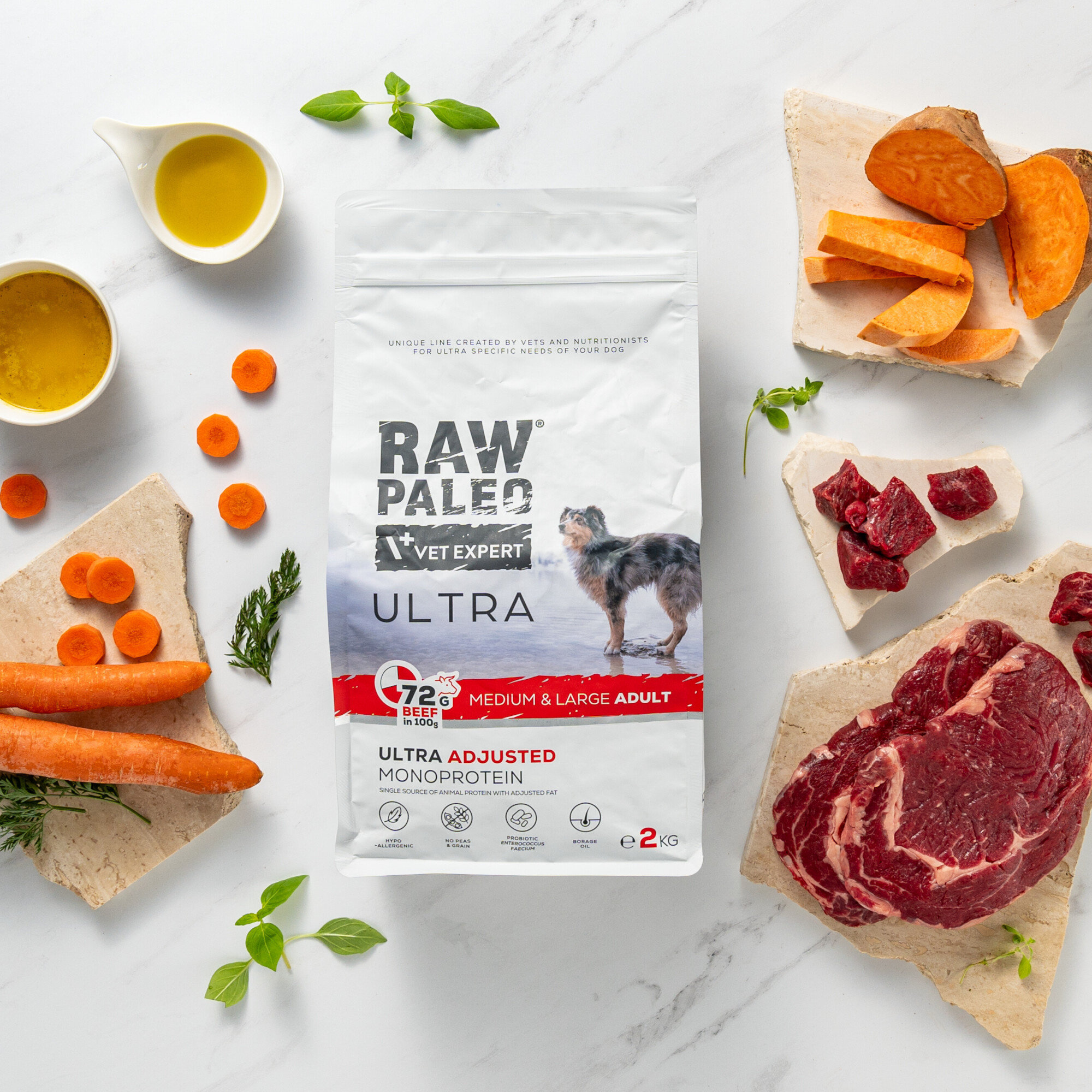 Na obrazie znajduje się opakowanie karmy dla psów \'RAW PALEO VET EXPERT ULTRA\' z wizerunkiem psa. Wokół opakowania leżą składniki takie jak marchewki, kawałki mięsa i słodkie ziemniaki.