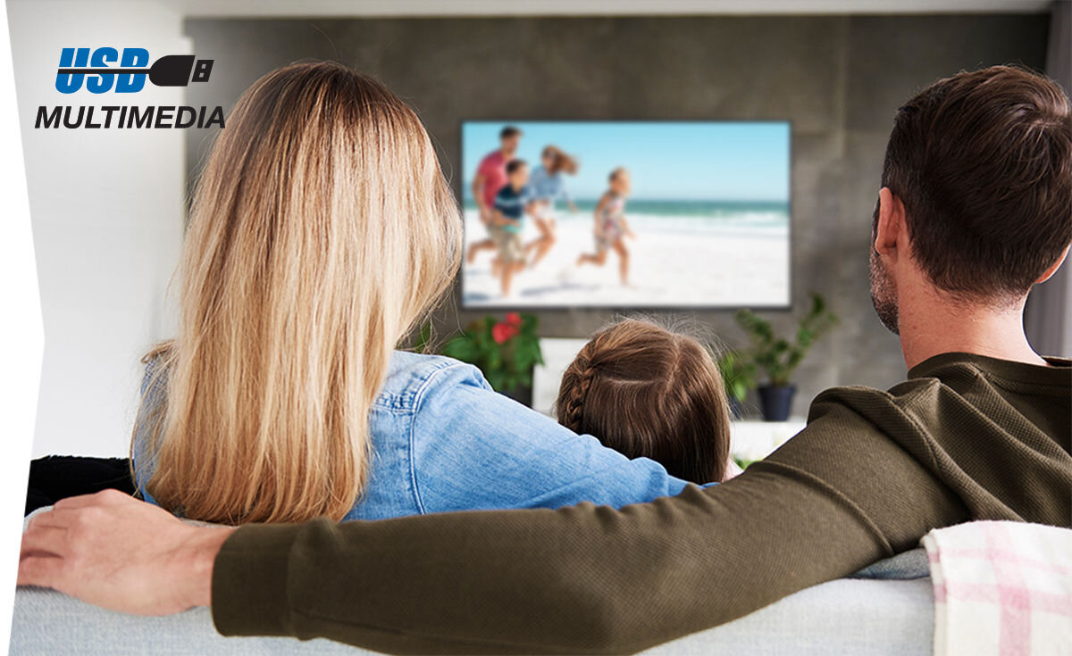 Trzy osoby siedzą na kanapie i oglądają telewizor, na którym wyświetlany jest obraz plaży z bawiącymi się dziećmi. W lewym górnym rogu widoczny jest napis \'USB MULTIMEDIA\'.