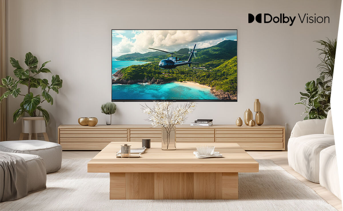 W salonie na ścianie wisi telewizor wyświetlający obraz helikoptera nad tropikalnym krajobrazem. W prawym górnym rogu ekranu widnieje napis \'Dolby Vision\'.