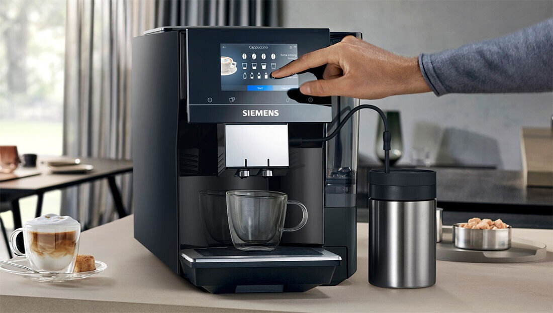 Czarny ekspres do kawy z logo SIEMENS, z dotykowym panelem sterowania, na którym widoczne są ikony różnych napojów. Obok ekspresu stoi metalowy pojemnik, a przed nim szklanka i filiżanka z kawą.