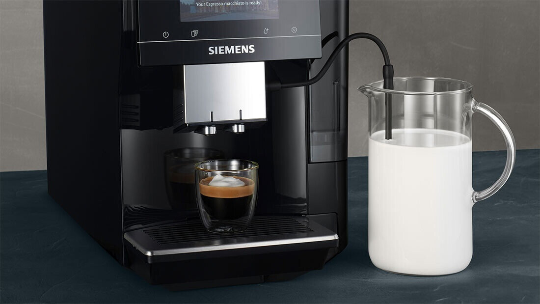 Czarny ekspres do kawy z logo SIEMENS, z filiżanką espresso na podstawce. Obok znajduje się szklany dzbanek z mlekiem podłączony rurką do ekspresu.