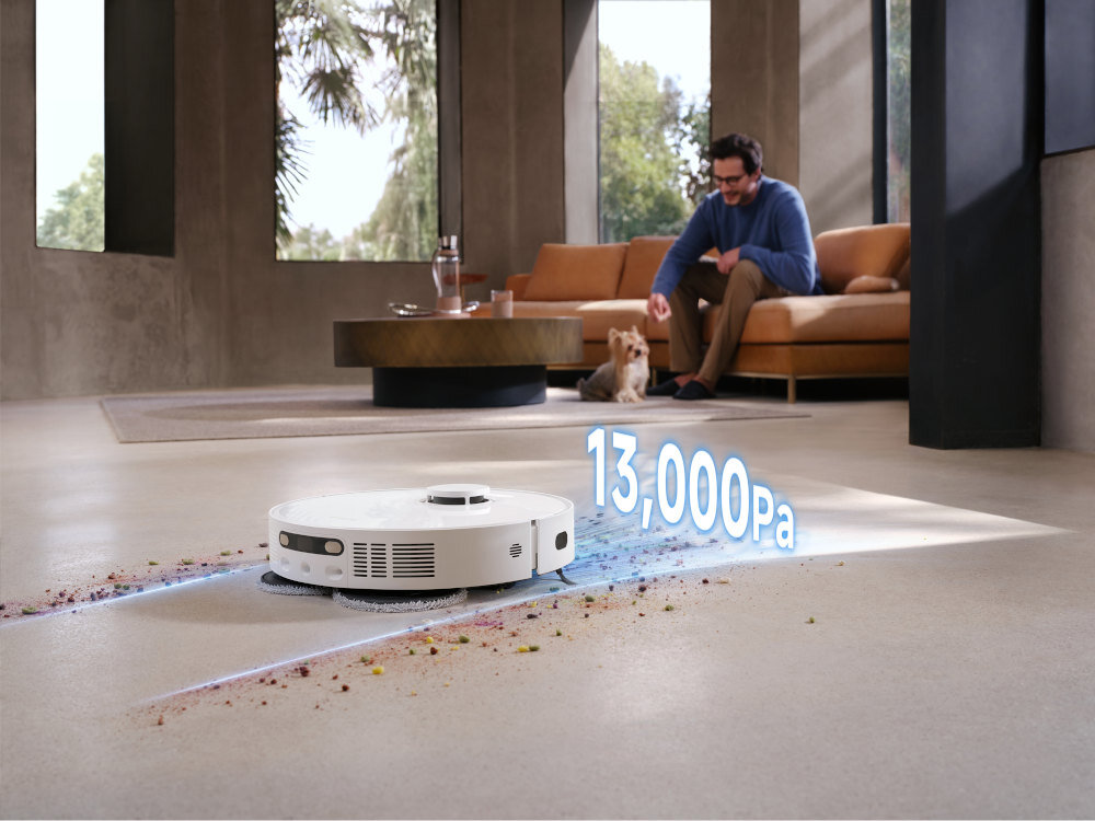 Na podłodze w nowoczesnym wnętrzu znajduje się biały robot sprzątający, obok którego widoczny jest napis \'13,000Pa\'. W tle siedzi mężczyzna na kanapie z psem.