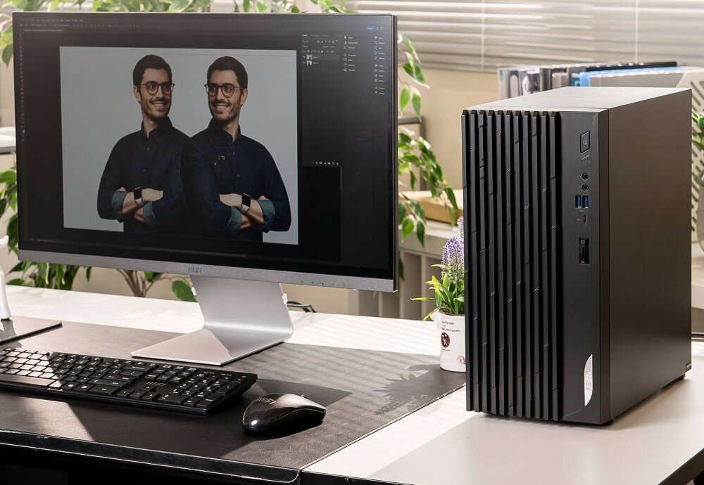 Na biurku znajduje się monitor z wyświetlonym zdjęciem dwóch mężczyzn oraz czarna obudowa komputera z widocznymi portami USB. W tle widać rośliny i książki na półkach.