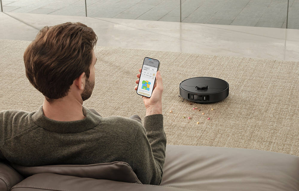 Osoba siedzi na kanapie, trzymając w ręku smartfon z aplikacją pokazującą mapę pomieszczenia. Na dywanie obok znajduje się czarny robot sprzątający, wokół którego leżą rozsypane drobinki.