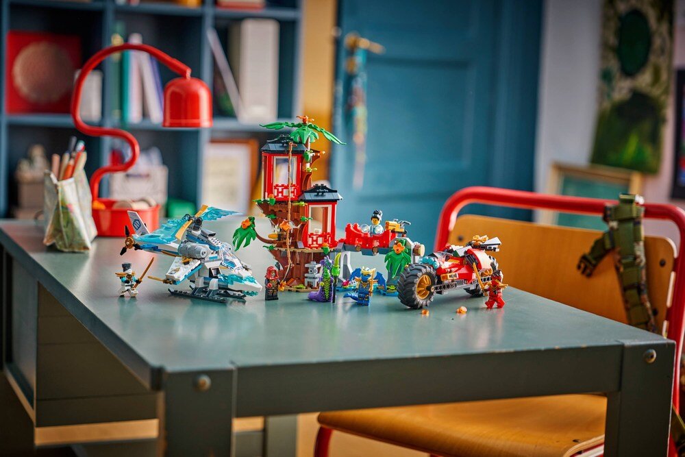 Na stole znajduje się zestaw klocków LEGO przedstawiający domek na drzewie z pojazdami i figurkami. W tle widać czerwoną lampkę i półkę z książkami.