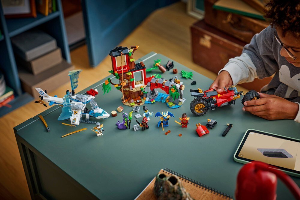 Na stole znajduje się zestaw klocków LEGO przedstawiający domek na drzewie z pojazdami i figurkami. Obok zestawu widoczna jest osoba składająca jeden z elementów.