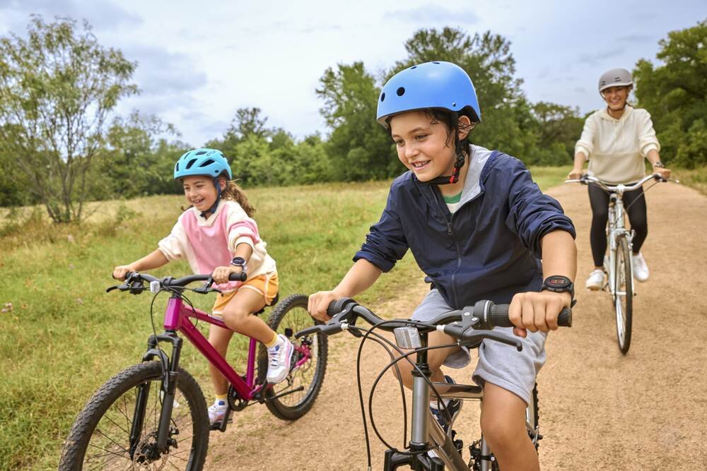 Trzy osoby jadą na rowerach po ścieżce w parku, dwie z nich mają na nadgarstkach zegarki sportowe. W tle widać drzewa i trawę.