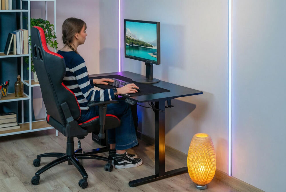 Osoba siedzi przy czarnym biurku elektrycznym z regulacją wysokości, na którym znajduje się monitor i klawiatura. Obok biurka stoi lampa podłogowa, a w tle widać regał z książkami i rośliną.