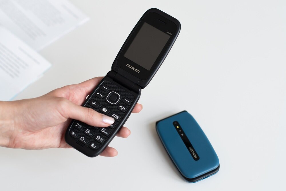 Osoba trzyma otwarty telefon z klapką z widocznym logo \'maxcom\' na ekranie. Obok leży zamknięty telefon w kolorze niebieskim z tym samym logo.