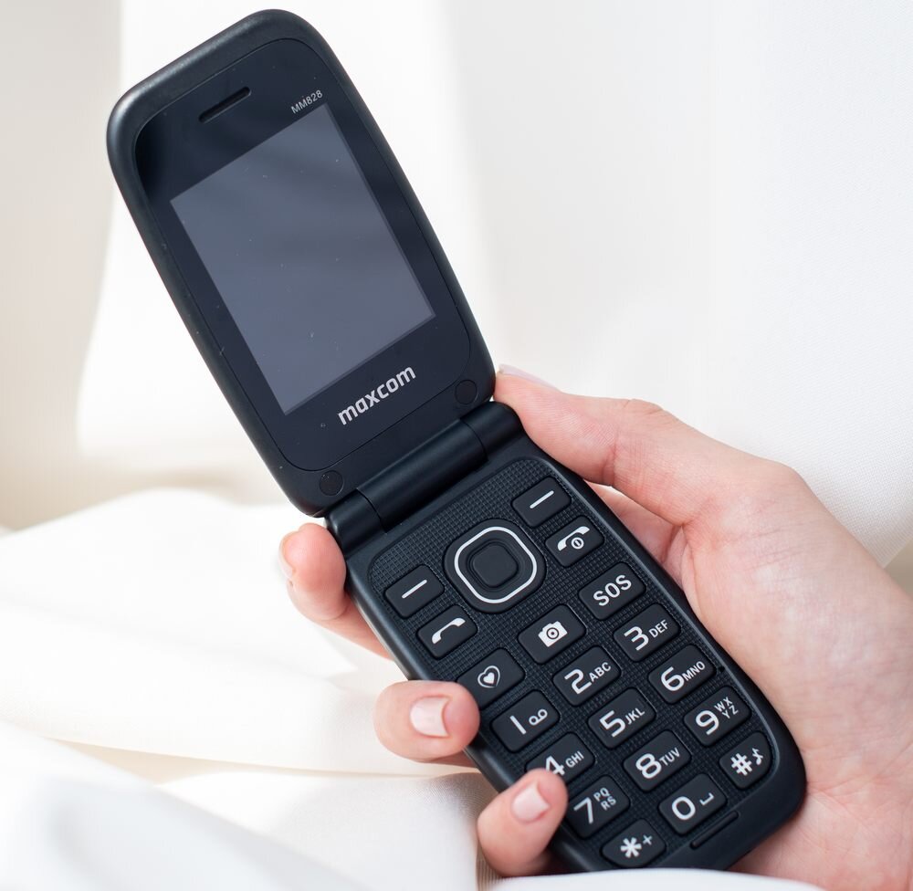 Osoba trzyma otwarty telefon z klapką marki maxcom, model MM 828. Na klawiaturze widoczne są duże przyciski z cyframi i symbolem SOS.